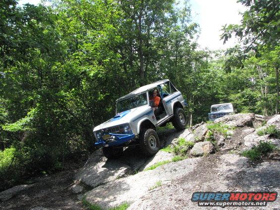White and blue Ford Bronco climbing a rocky hill, another Bronco in background