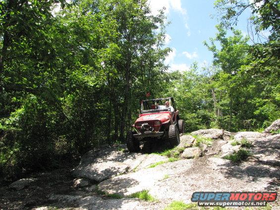 Red Jeep Wrangler driving over rocks on a wooded trail