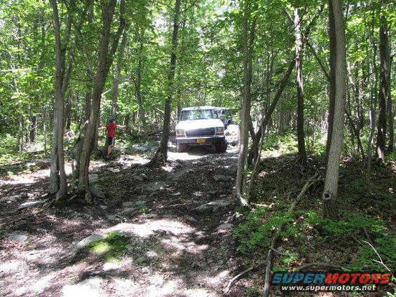 Two SUVs navigate a rocky, wooded trail. One vehicle is in the foreground, the other behind it