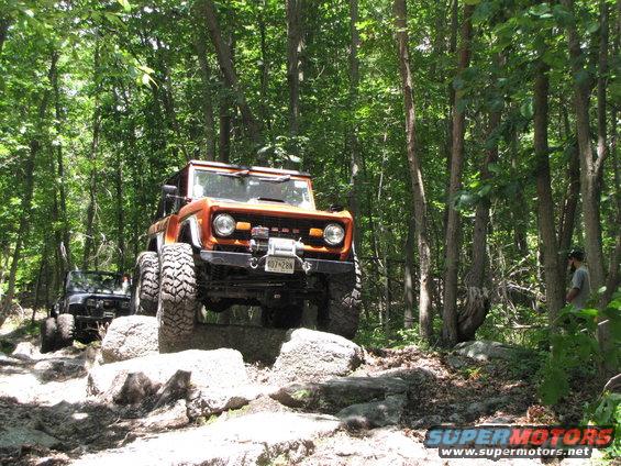 orange 1979 Ford Bronco climbing rocks on wooded trail, another SUV behind it
