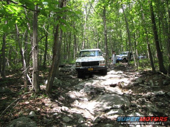 White 1987 Ford Bronco on a rocky forest trail with another SUV behind it