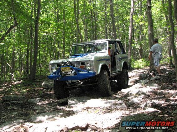 Silver Ford Bronco with blue bumper and roll cage driving on a rocky forest trail