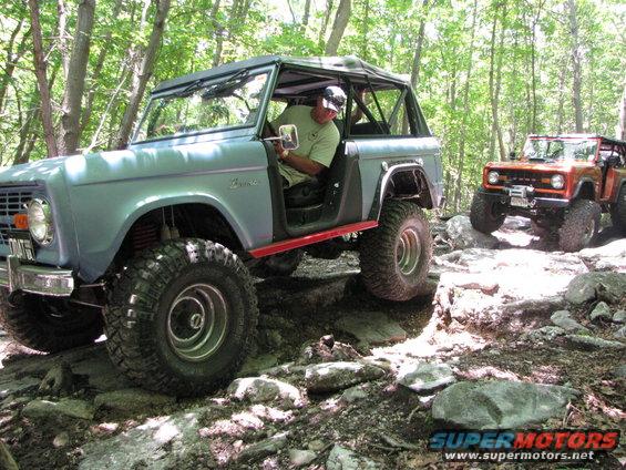 Light blue 1978 Ford Bronco with red trim, driven off-road on rocks. Orange Bronco follows