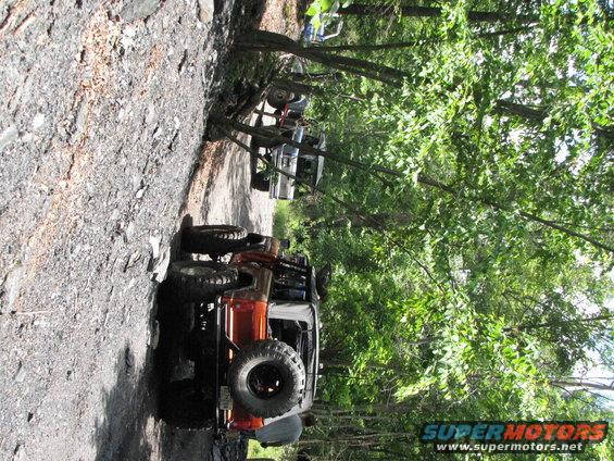 Several off-road vehicles parked on a wooded dirt path, one in the foreground is orange and brown with a spare tire on the back