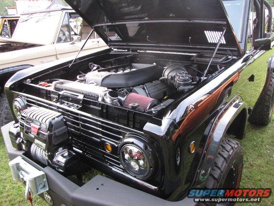 Black Ford Bronco with hood open, showing engine and winch. Another Bronco is visible in the background