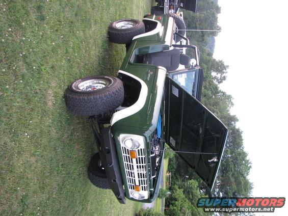 Green Ford Bronco with white stripe, hood open, parked on grass. Another vehicle is visible behind it