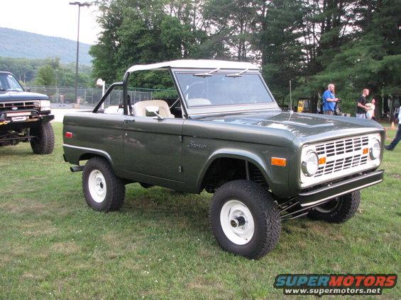 Green 1978 Ford Bronco with a white soft top, parked on grass next to a dark pickup truck