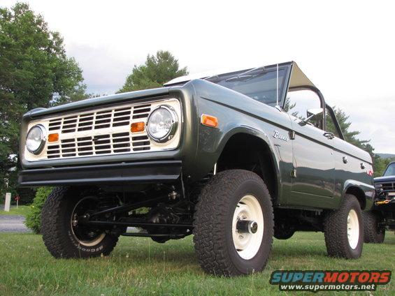 Green Ford Bronco with white grille and white wheels, parked on grass. Another vehicle is partially visible to the right