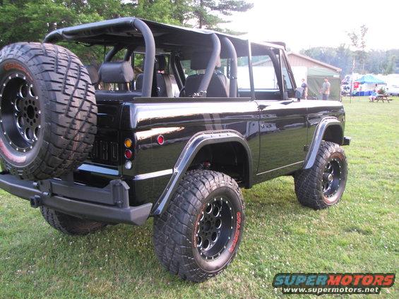 Black Ford Bronco, lifted with large tires, spare tire mounted on rear swing away carrier. Roll cage visible inside. Site has tents and picnic tables in background