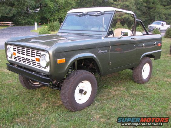 Green Ford Bronco with white top parked on grass, white pickup truck in background