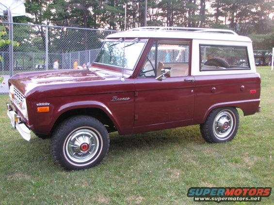 Maroon Ford Bronco with white hardtop parked on grass. Bronco badge on fender