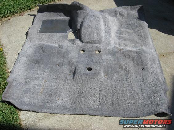 Gray car carpet laid out on concrete, showing floor mat area and bolt holes