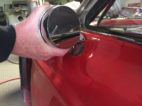 Hand holding a round chrome side mirror against the red door of a car. Another car body is visible in the background