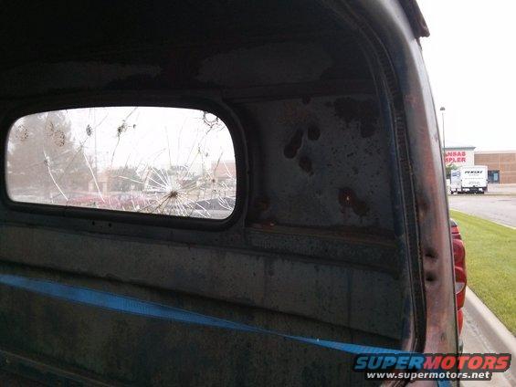 Interior view of a truck cab showing a cracked rear window and rusted metal panels. A blue strap is visible