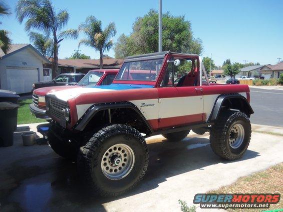 Red, white, and blue Bronco with lifted suspension and off-road tires parked in a driveway. Another vehicle is visible behind it