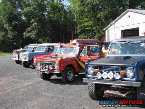 Four Ford Broncos parked in a line, front one is blue, next is orange, then red, then blue