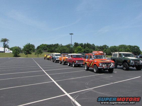 Line of Ford Broncos parked in a lot, front-most is orange with yellow stripes and roof rack