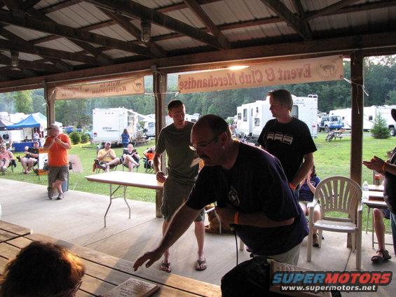 People gathered under a covered pavilion at an outdoor event with RVs and tents in the background