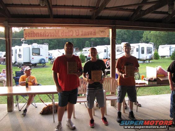 Three men stand holding plaques under a covered patio, with RVs parked in the background