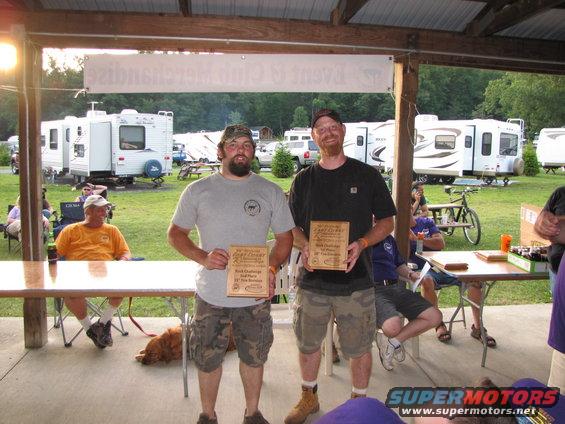 Two men hold wooden awards at an outdoor gathering; RVs and trees are visible in the background