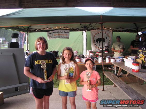 Three girls with trophies at an outdoor event with tents and tables