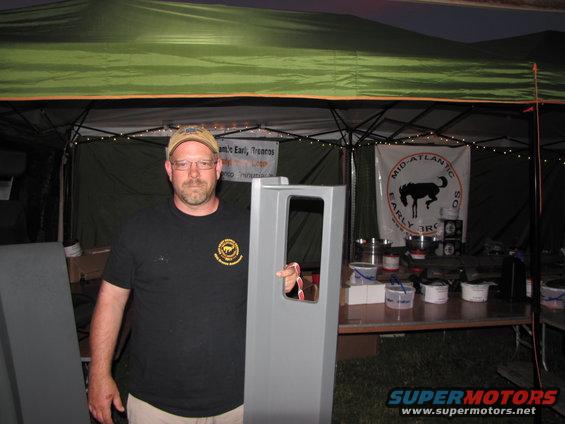 Man in cap and t-shirt holds gray car door in front of tent with banners