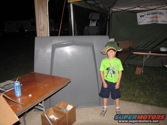 Kid wearing a big hat stands in front of a gray car hood