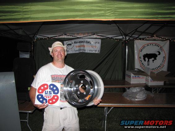 Man in hat and t-shirt holds a car wheel and a US Mags sticker. Banner behind him says Mid-Atlantic Early Broncos