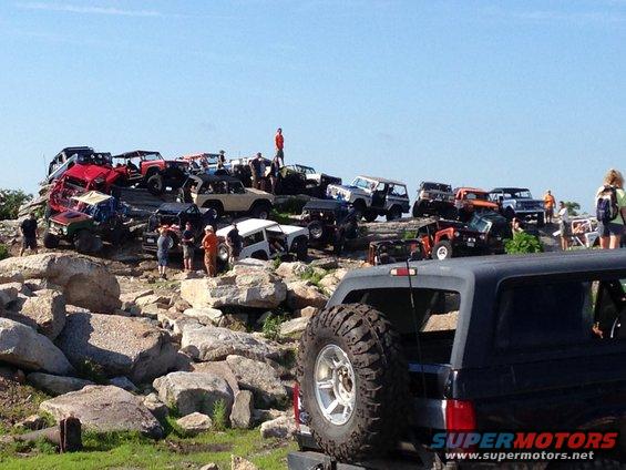 Many off-road vehicles parked on a rocky hill under a clear blue sky. A few people are standing around the vehicles