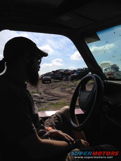 Man with beard and cap looks out car window at a junkyard full of old vehicles