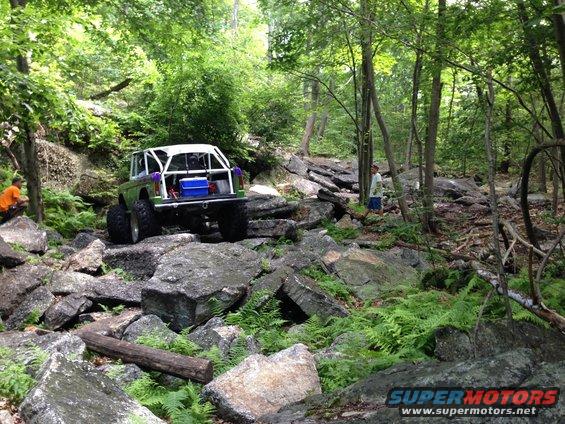 Green Ford Bronco with white roll cage and blue cooler on rear, navigating a rocky trail