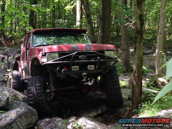 Red truck with off-road tires navigating rocks in a forest. Has black racing stripes on the hood and a custom front bumper with PIAA lights