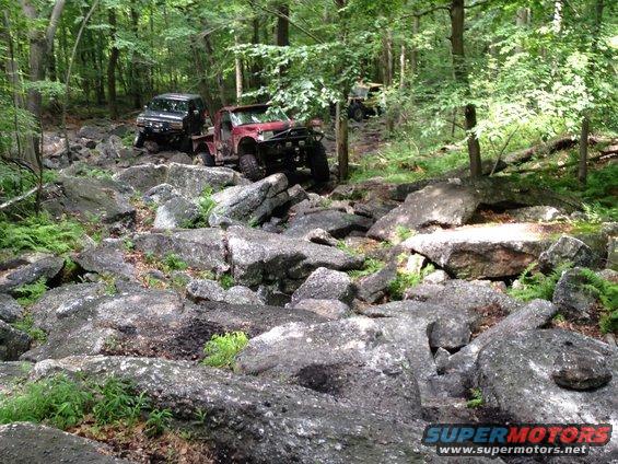 Two off-road trucks and an ATV on a rocky forest trail