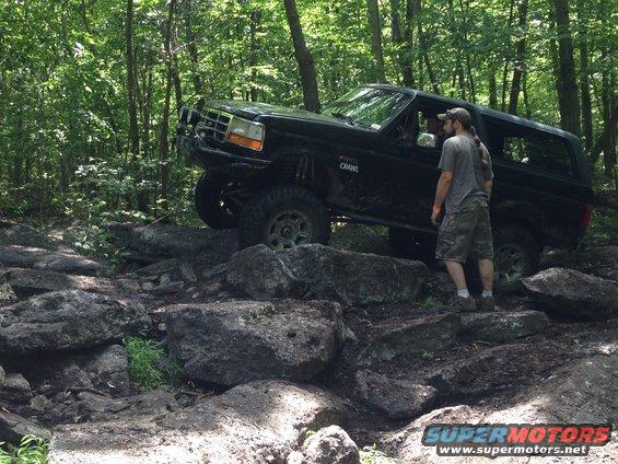 Black Ford Bronco navigating a rocky trail in the woods. A man stands nearby