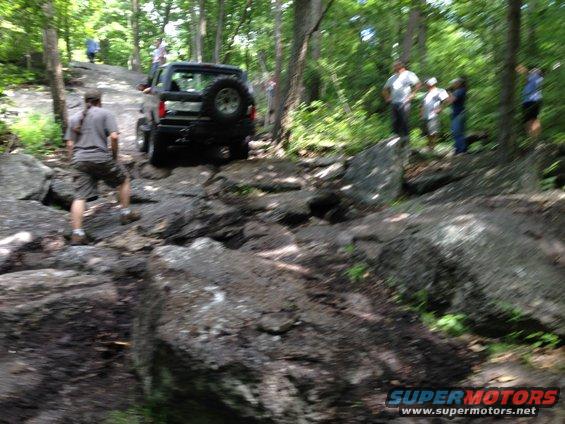 Black SUV with spare tire on rear door navigating a rocky trail with people watching