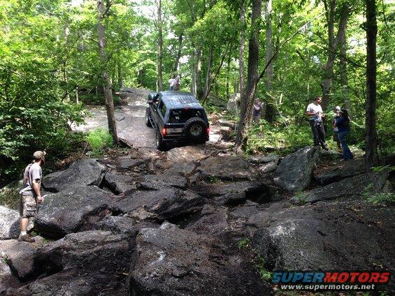 Black SUV driving over large rocks on a wooded trail. People watch from the side