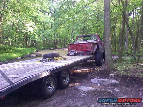 Red pickup truck on a trailer in a wooded area near a stream. Truck has 'FORD' on tailgate