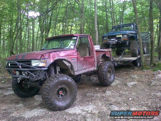 Red pickup truck with large tires and off-road modifications, towing a blue off-road vehicle on a trailer
