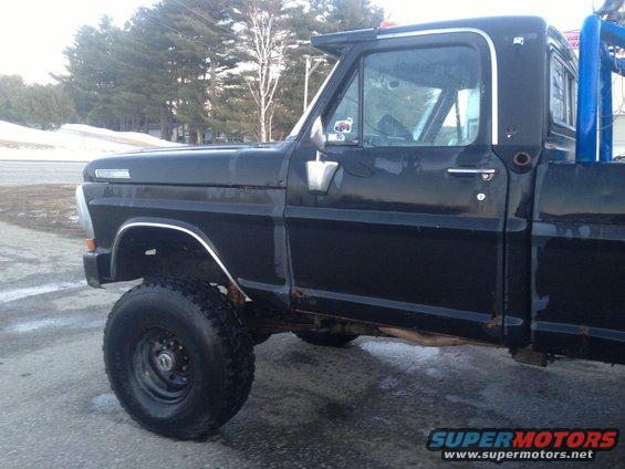 Black 1970 Ford F-250 pickup truck, side view, showing rust on the lower body panels and a blue roll bar in the bed