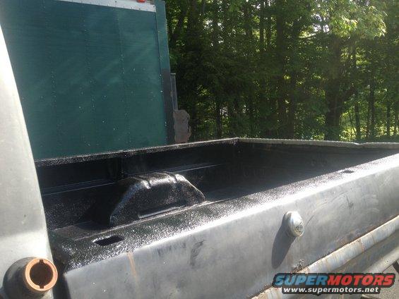 Truck bed with black liner, side view. A green trailer is behind it