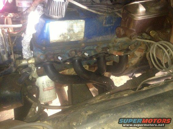Engine bay view showing a V8 engine with exhaust manifolds, spark plugs, and an oil filter. Text on oil filter is visible