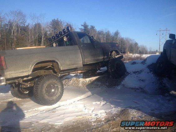 Lifted pickup truck with dual rear wheels driving off-road in snow. Another truck is visible to the right