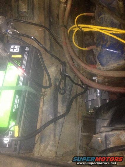 Battery and alternator in a vehicle engine bay. The battery is green and black with positive terminal marked