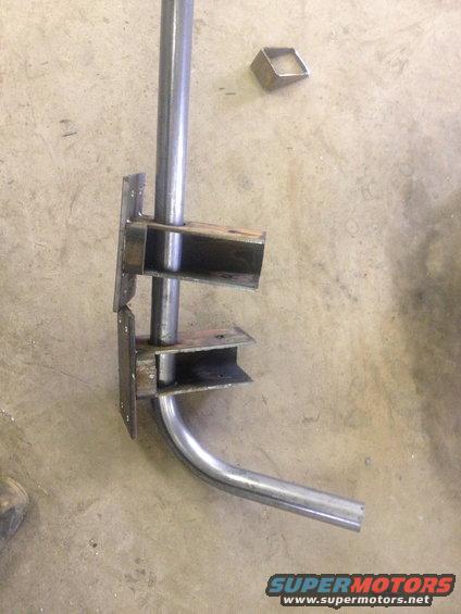 Metal pipe bent and welded to brackets, lying on concrete floor. Small metal square nearby