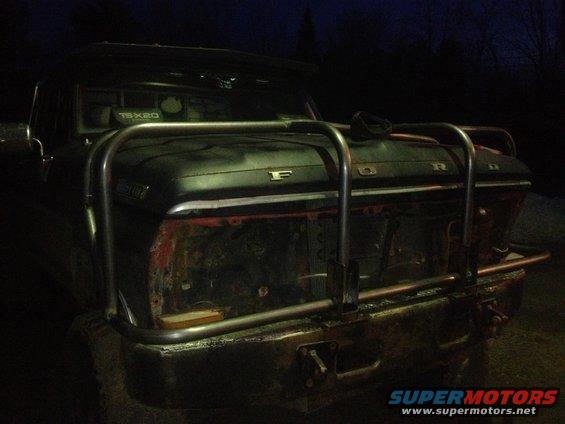 1970 Ford F-250 front end with custom bull bar and missing grille. Dark exterior, red inner fender