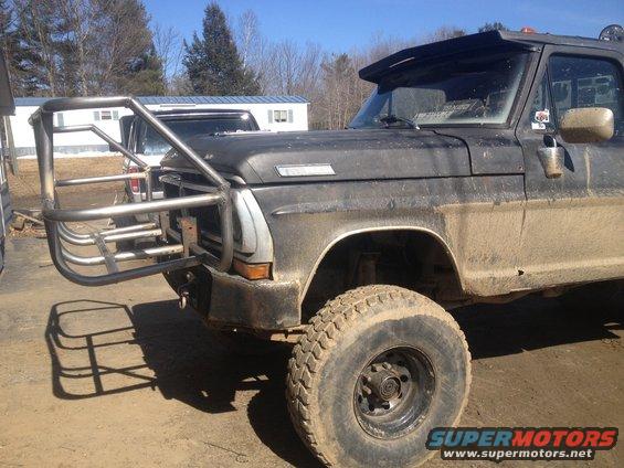 Front quarter view of a dark-colored Ford pickup truck with a large, custom-made metal bumper and brush guard. The truck appears dirty and has large off-road tires