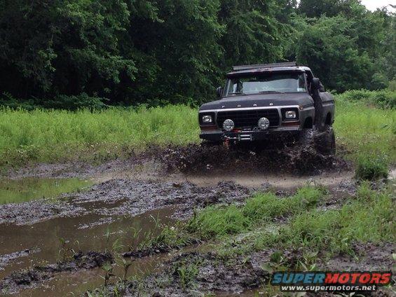 Black 1979 Ford Bronco driving through mud, kicking up spray. It has a roof rack and off-road lights