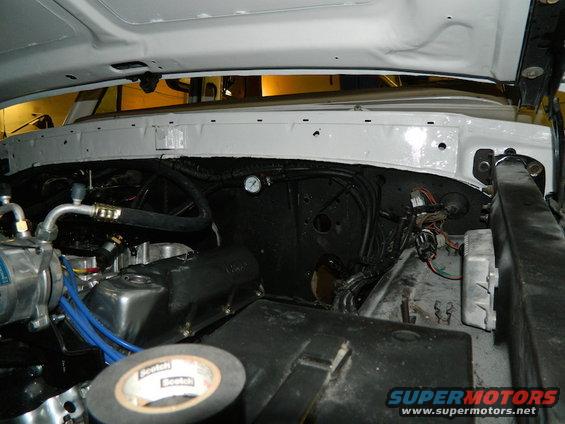 White vehicle engine bay with black firewall, hoses, wiring, and a gauge. Scotch tape roll in foreground