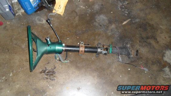 Green steering column with shifter lever and wiring harness detached, lying on a concrete floor