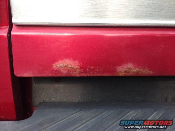 Red vehicle tailgate with visible rust spots on the lower panel and a silver trim piece above